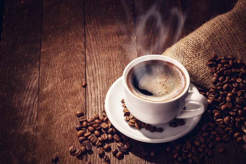 A steaming cup of coffee with roasted beans in a burlap sack